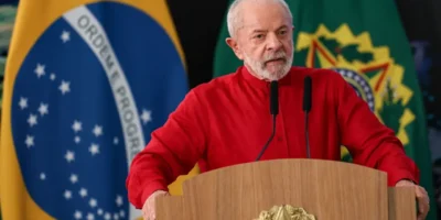 Presidente Lula, de camisa vermelha, discursa diante de dois microfones, com a bandeira brasileira e a bandeira presidencial ao fundo.