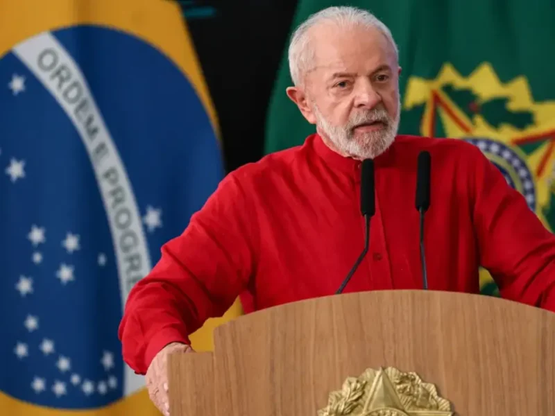 Presidente Lula, de camisa vermelha, discursa diante de dois microfones, com a bandeira brasileira e a bandeira presidencial ao fundo.