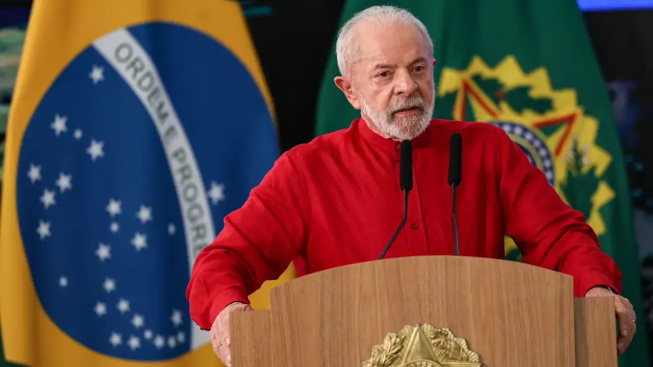 Presidente Lula, de camisa vermelha, discursa diante de dois microfones, com a bandeira brasileira e a bandeira presidencial ao fundo. Presidente Lula, de camisa vermelha, discursa diante de dois microfones, com a bandeira brasileira e a bandeira presidencial ao fundo.
