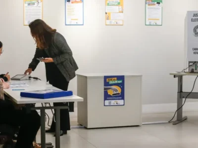Pessoas realizam procedimentos em uma seção eleitoral, com urna eletrônica instalada ao lado da mesa dos mesários.