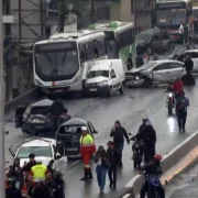 Vista geral do engavetamento na Via Dutra mostra ônibus, carros destruídos, equipes de resgate e pessoas caminhando entre os veículos acidentados.