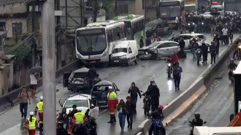 Vista geral do engavetamento na Via Dutra mostra ônibus, carros destruídos, equipes de resgate e pessoas caminhando entre os veículos acidentados.