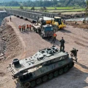 Blindado militar, caminhão e equipes de trabalhadores próximos à montagem de uma ponte metálica sobre um rio.