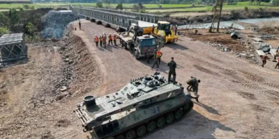 Blindado militar, caminhão e equipes de trabalhadores próximos à montagem de uma ponte metálica sobre um rio.