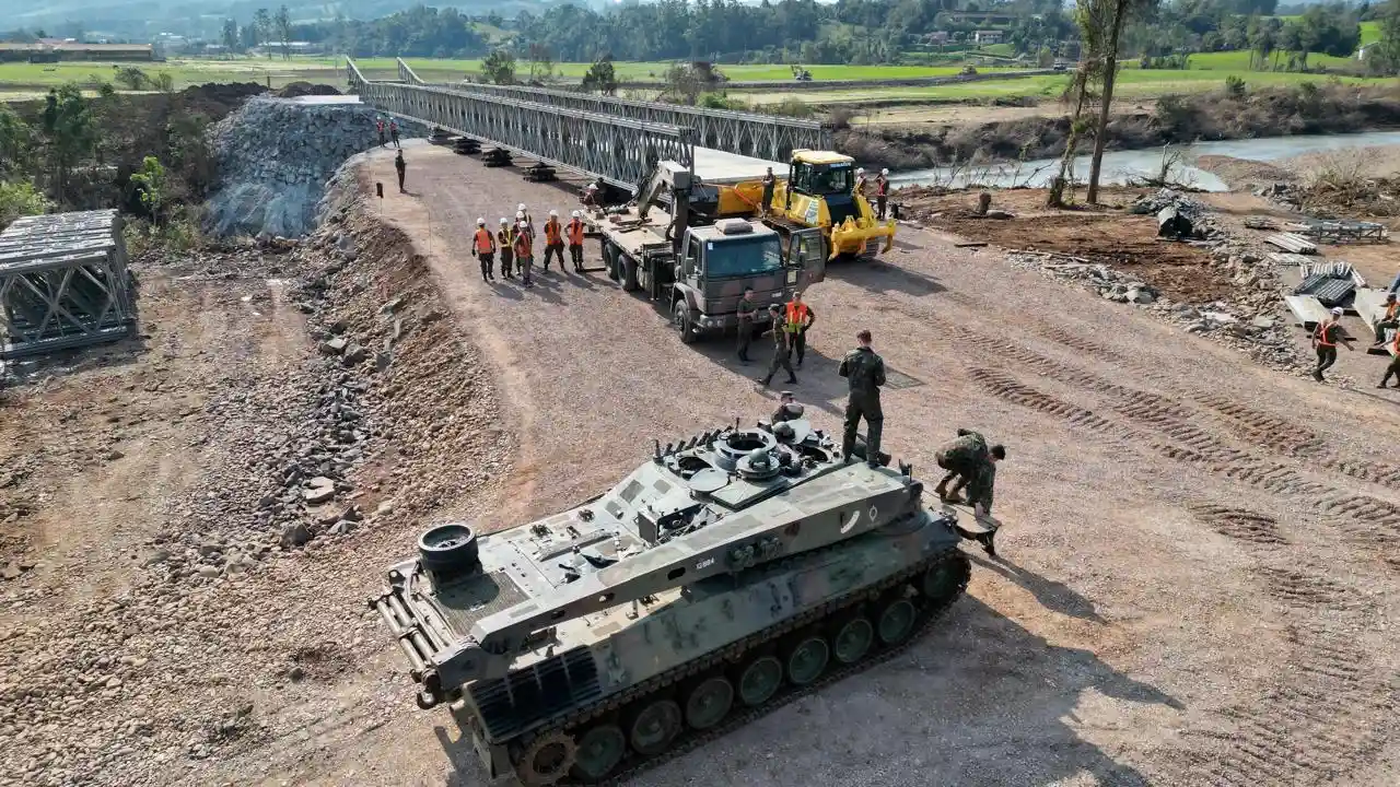 Blindado militar, caminhão e equipes de trabalhadores próximos à montagem de uma ponte metálica sobre um rio. Blindado militar, caminhão e equipes de trabalhadores próximos à montagem de uma ponte metálica sobre um rio.