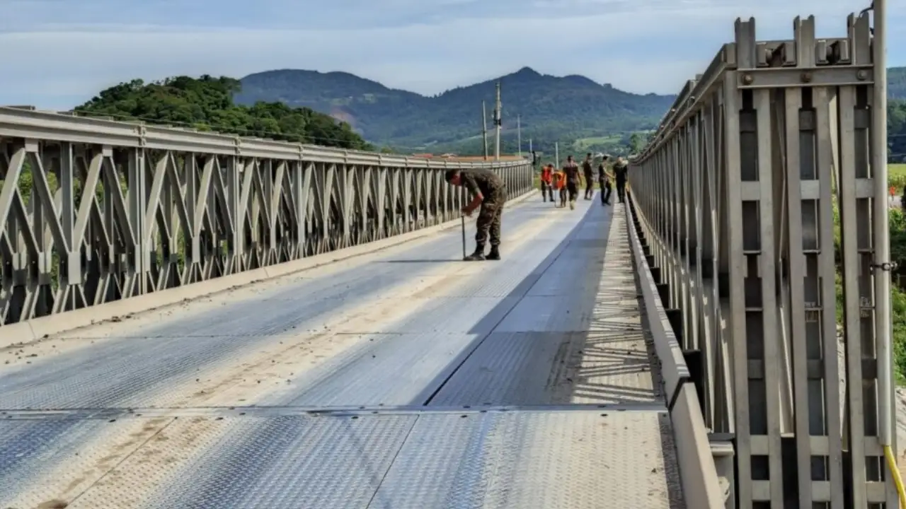Militar inspeciona o piso de uma ponte metálica modular, enquanto outras pessoas caminham pela estrutura ao fundo. Militar inspeciona o piso de uma ponte metálica modular, enquanto outras pessoas caminham pela estrutura ao fundo.