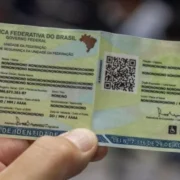 Mão segura modelo ilustrativo de carteira de identidade brasileira, com QR Code, fotografia representativa e dados pessoais fictícios.