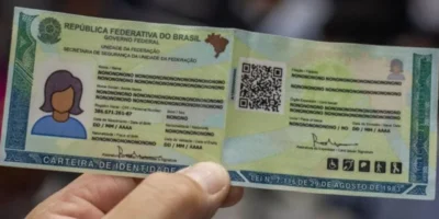 Mão segura modelo ilustrativo de carteira de identidade brasileira, com QR Code, fotografia representativa e dados pessoais fictícios.