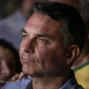 Senador Flávio Bolsonaro aparece em primeiro plano, com expressão séria, durante evento público, cercado por apoiadores.