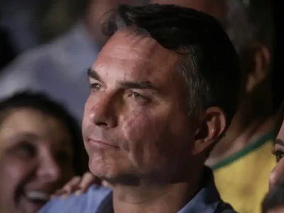 Senador Flávio Bolsonaro aparece em primeiro plano, com expressão séria, durante evento público, cercado por apoiadores.