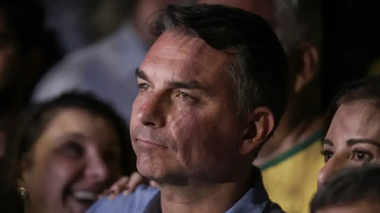 Senador Flávio Bolsonaro aparece em primeiro plano, com expressão séria, durante evento público, cercado por apoiadores.