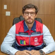Haroldinho Jesus sentado à mesa, usando óculos e colete vermelho e azul com identificação da Prefeitura de Itaguaí.