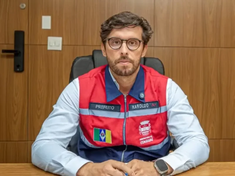 Haroldinho Jesus sentado à mesa, usando óculos e colete vermelho e azul com identificação da Prefeitura de Itaguaí.