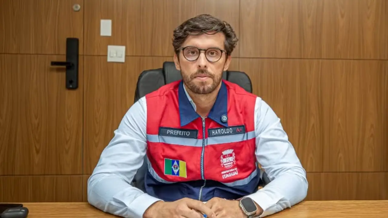 Haroldinho Jesus sentado à mesa, usando óculos e colete vermelho e azul com identificação da Prefeitura de Itaguaí.