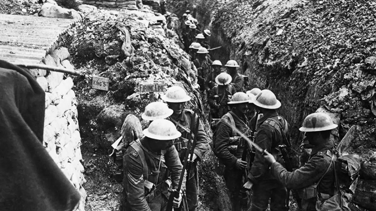 Soldados britânicos em uma trincheira enlameada na Bélgica, durante a Primeira Guerra Mundial, cercados por sacos de areia e equipamentos militares.