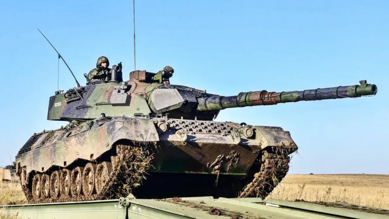Carro de combate Leopard 1A5 BR camuflado, visto de lado, com militares na torre durante passagem sobre uma estrutura metálica. Carro de combate Leopard 1A5 BR camuflado, visto de lado, com militares na torre durante passagem sobre uma estrutura metálica.