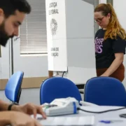 Eleitora vota em cabine da Justiça Eleitoral enquanto mesário trabalha em seção eleitoral.