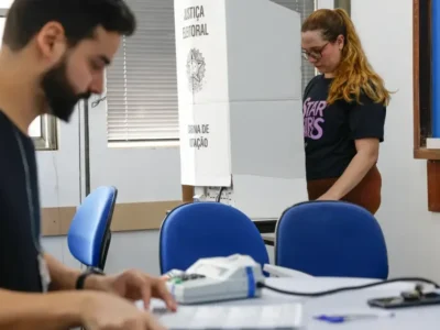 Eleitora vota em cabine da Justiça Eleitoral enquanto mesário trabalha em seção eleitoral.