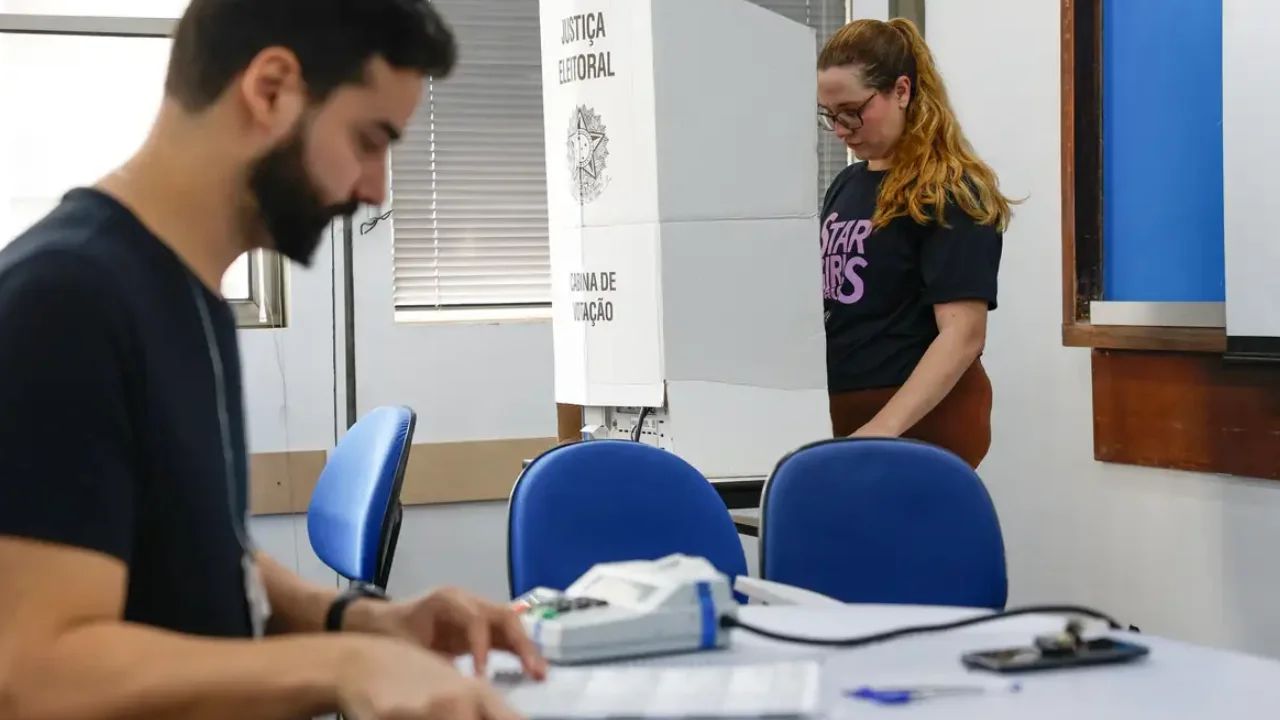 Eleitora vota em cabine da Justiça Eleitoral enquanto mesário trabalha em seção eleitoral.