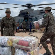 Militares do Exército Brasileiro movimentam suprimentos embalados ao lado de um helicóptero militar em uma área de pouso.
