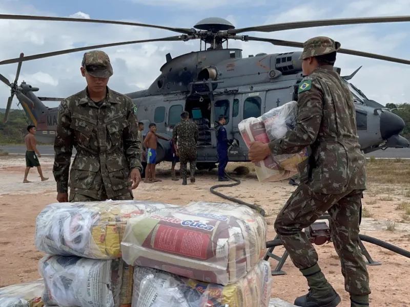 Militares do Exército Brasileiro movimentam suprimentos embalados ao lado de um helicóptero militar em uma área de pouso.