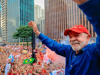 Presidente Lula, de boné vermelho e camisa azul, acena com os braços levantados para uma multidão de apoiadores com bandeiras.