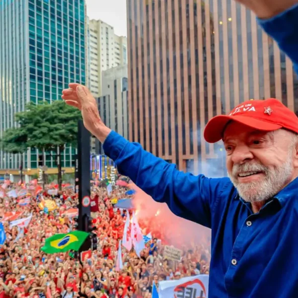 Presidente Lula, de boné vermelho e camisa azul, acena com os braços levantados para uma multidão de apoiadores com bandeiras.
