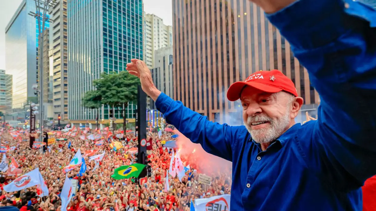 Presidente Lula, de boné vermelho e camisa azul, acena com os braços levantados para uma multidão de apoiadores com bandeiras. Presidente Lula, de boné vermelho e camisa azul, acena com os braços levantados para uma multidão de apoiadores com bandeiras.