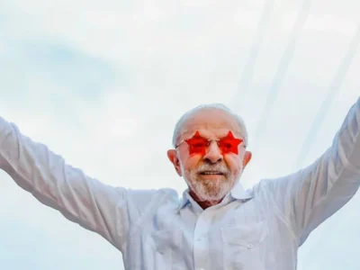 Presidente Lula usa óculos vermelhos em formato de estrelas e levanta os dois braços com os polegares apontados para cima.