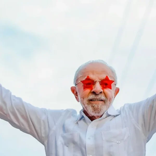Presidente Lula usa óculos vermelhos em formato de estrelas e levanta os dois braços com os polegares apontados para cima.