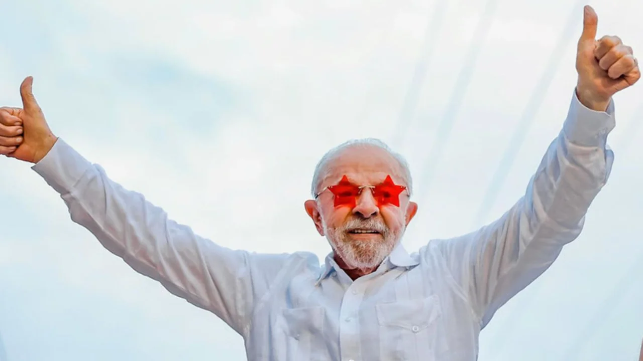 Presidente Lula usa óculos vermelhos em formato de estrelas e levanta os dois braços com os polegares apontados para cima. Presidente Lula usa óculos vermelhos em formato de estrelas e levanta os dois braços com os polegares apontados para cima.