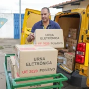 Agente de segurança armado acompanha o descarregamento de urnas eletrônicas de um veículo amarelo.