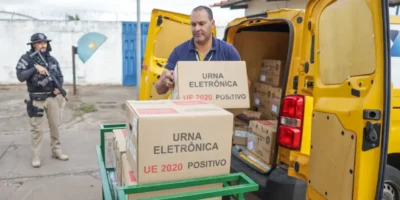 Agente de segurança armado acompanha o descarregamento de urnas eletrônicas de um veículo amarelo.