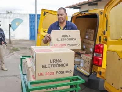 Agente de segurança armado acompanha o descarregamento de urnas eletrônicas de um veículo amarelo.