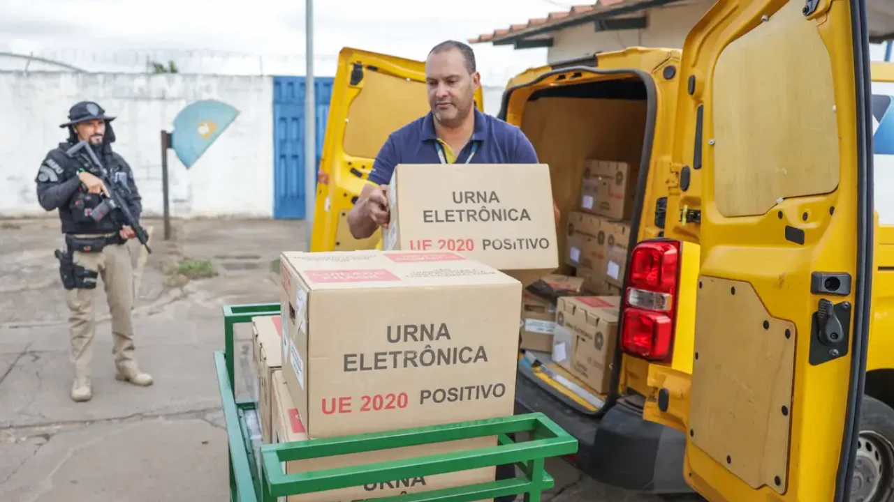 Agente de segurança armado acompanha o descarregamento de urnas eletrônicas de um veículo amarelo.