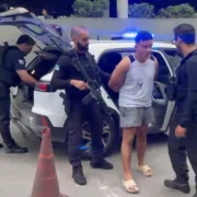 Policiais armados conduzem um homem ao lado de uma viatura com luzes de emergência azuis acionadas.