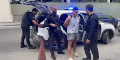 Policiais armados conduzem um homem ao lado de uma viatura com luzes de emergência azuis acionadas.