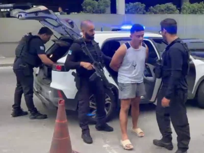 Policiais armados conduzem um homem ao lado de uma viatura com luzes de emergência azuis acionadas.
