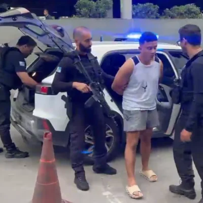 Policiais armados conduzem um homem ao lado de uma viatura com luzes de emergência azuis acionadas.