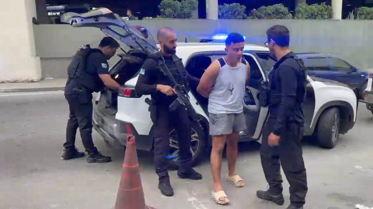 Policiais armados conduzem um homem ao lado de uma viatura com luzes de emergência azuis acionadas.