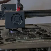 Impressora 3D em funcionamento, com cabeçote depositando plástico sobre uma peça em fabricação.