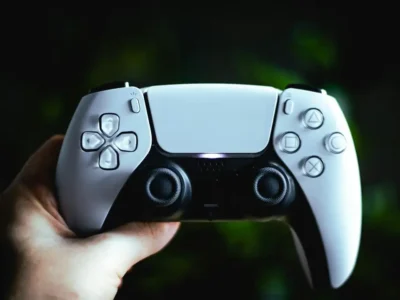 Mão segura controle DualSense branco e preto do PlayStation 5 diante de um fundo escuro.