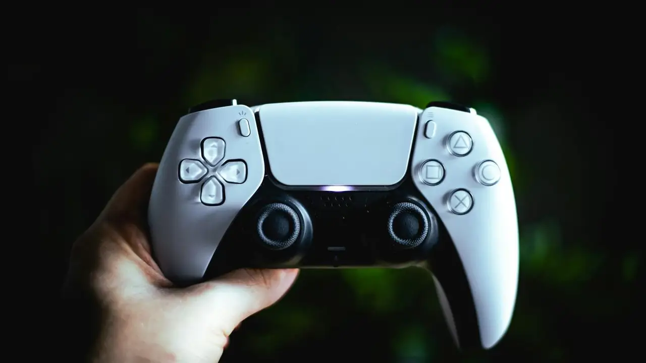Mão segura controle DualSense branco e preto do PlayStation 5 diante de um fundo escuro.
