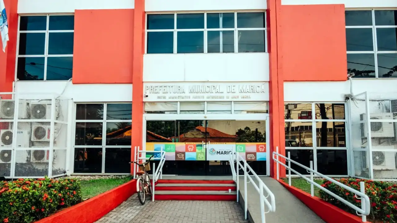 Fachada da Prefeitura Municipal de Maricá, com arquitetura em tons de vermelho e branco, jardins e calçada arborizada. Fachada da Prefeitura Municipal de Maricá, com arquitetura em tons de vermelho e branco, jardins e calçada arborizada.
