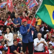Presidente Lula segura uma grande bandeira do Brasil durante ato político, cercado por uma multidão de apoiadores com bandeiras e cartazes.