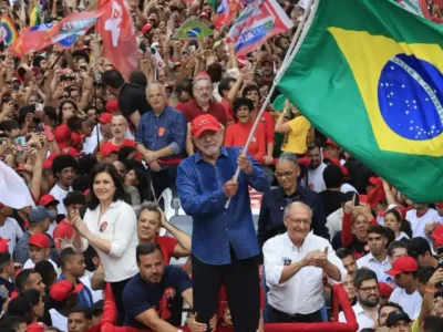 Presidente Lula segura uma grande bandeira do Brasil durante ato político, cercado por uma multidão de apoiadores com bandeiras e cartazes.
