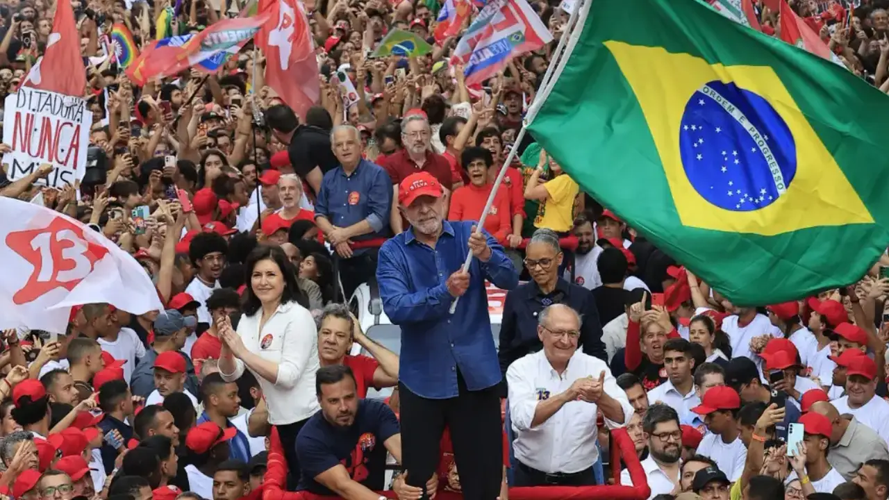Presidente Lula segura uma grande bandeira do Brasil durante ato político, cercado por uma multidão de apoiadores com bandeiras e cartazes.