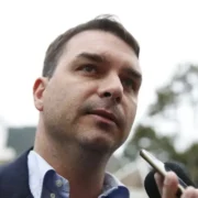 Senador Flávio Bolsonaro, de paletó escuro e camisa azul-clara, fala com jornalistas enquanto um celular é aproximado de seu rosto.