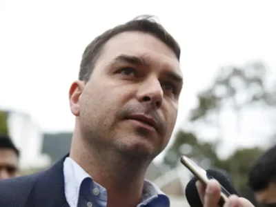 Senador Flávio Bolsonaro, de paletó escuro e camisa azul-clara, fala com jornalistas enquanto um celular é aproximado de seu rosto.