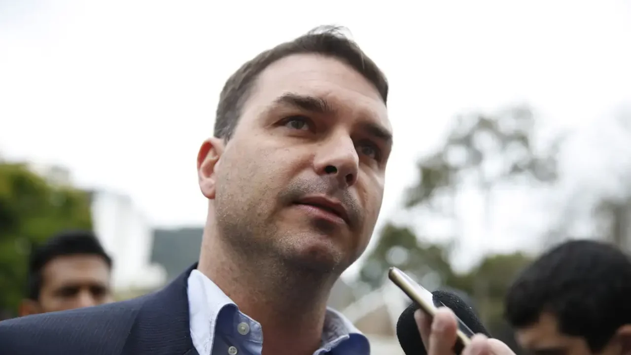 Senador Flávio Bolsonaro, de paletó escuro e camisa azul-clara, fala com jornalistas enquanto um celular é aproximado de seu rosto. Senador Flávio Bolsonaro, de paletó escuro e camisa azul-clara, fala com jornalistas enquanto um celular é aproximado de seu rosto.